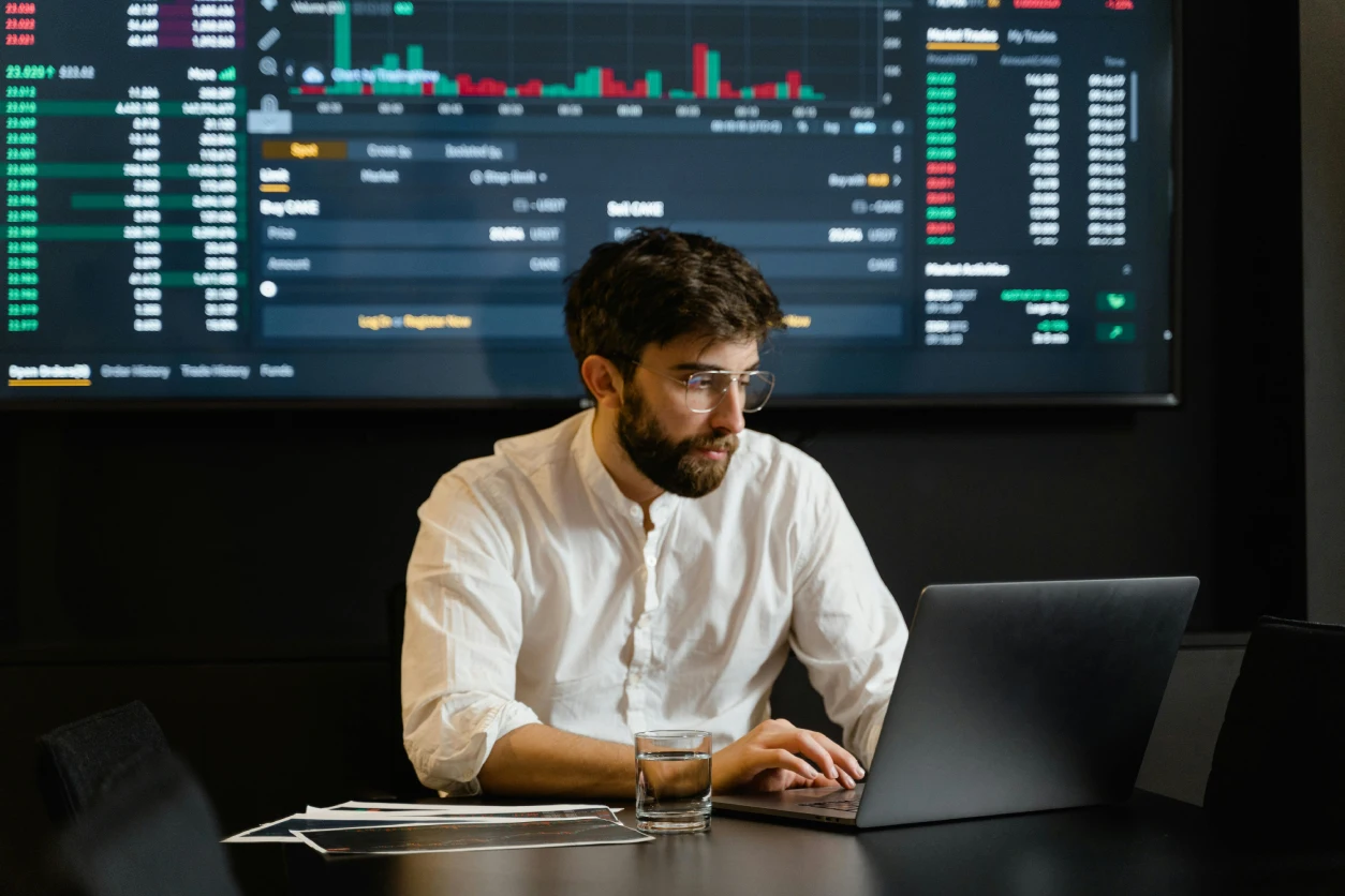 man sitting at a desk looking at a computer, A board of data is behind him.